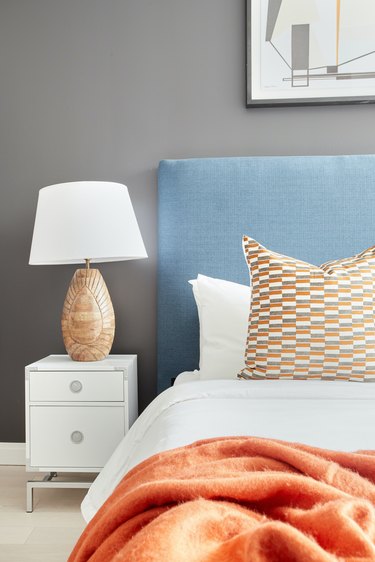 Eclectic bedroom with gray wall, blue upholstered headboard, lamp, white nightstand, orange blanket, pillow.